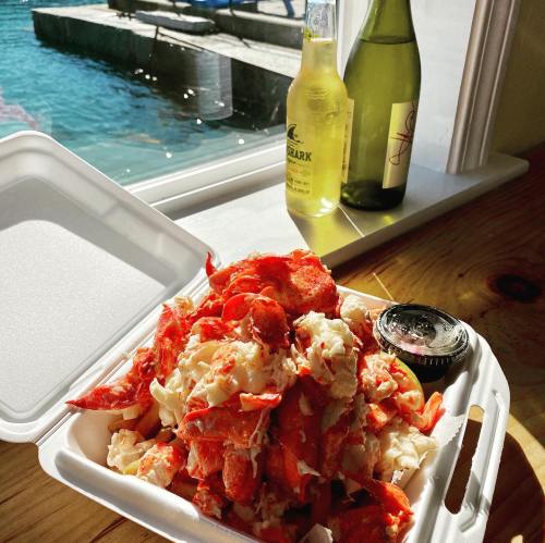 A takeout container filled with lobster meat, accompanied by a small sauce, with drinks and a waterfront view in the background.