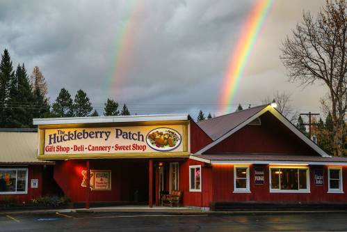 A red building with a sign reading "The Huckleberry Patch" under a cloudy sky with two rainbows in the background.