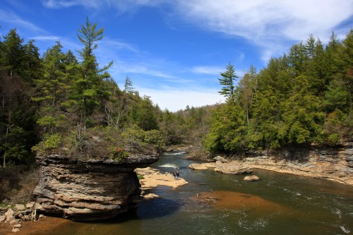 A serene river flows through a forested landscape, with rocky outcrops and clear blue skies above.