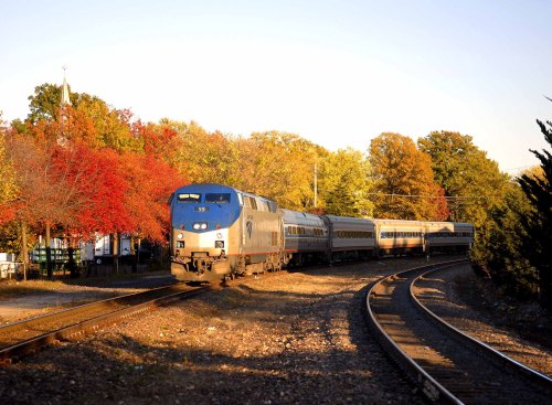 A blue and silver train curves around a track surrounded by vibrant autumn foliage.