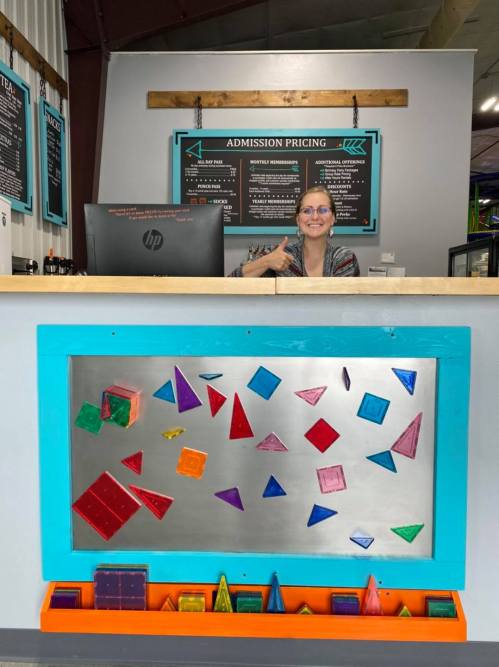 A smiling woman at a reception desk with colorful geometric shapes displayed on the wall behind her.