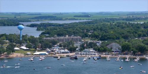 Aerial view of a lakeside area with boats, a water tower, and green trees under a clear blue sky.