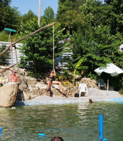 A person swings from a rope over a pool, while others relax on the shore and in the water surrounded by greenery.