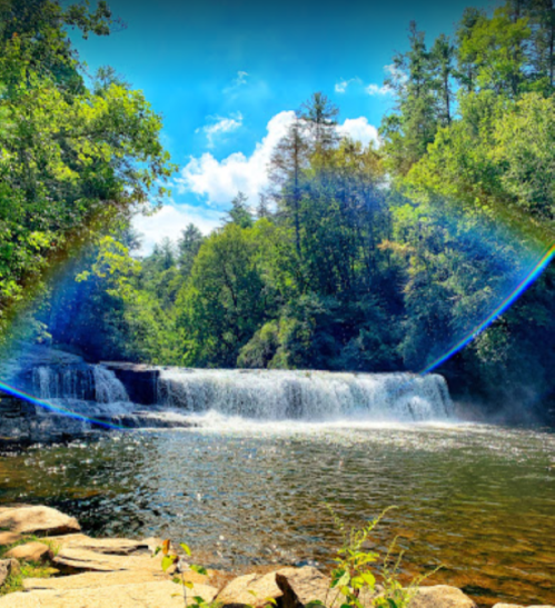 A serene waterfall surrounded by lush greenery and a clear blue sky, with a rainbow arching over the scene.
