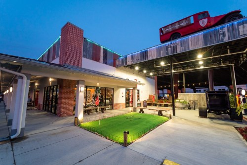 A modern building with a red vintage car on the roof, featuring outdoor seating and a green lawn area at dusk.