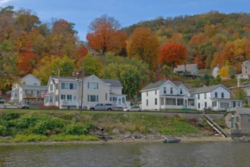 A scenic view of houses along a river, surrounded by vibrant autumn foliage on a hillside.
