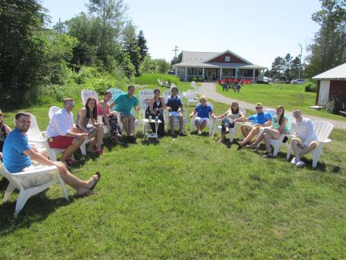 A group of people sitting in white chairs on a grassy lawn, enjoying a sunny day near a building in the background.