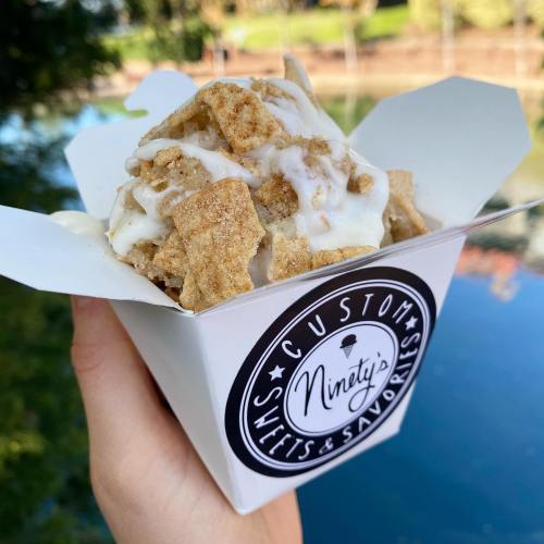 A hand holding a white takeout box filled with ice cream topped with crushed cereal and drizzled with frosting.