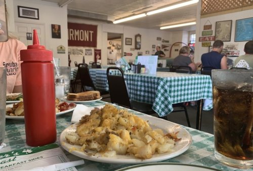 A plate of cheesy potatoes on a checkered table, with a ketchup bottle and a glass of cola in a diner setting.