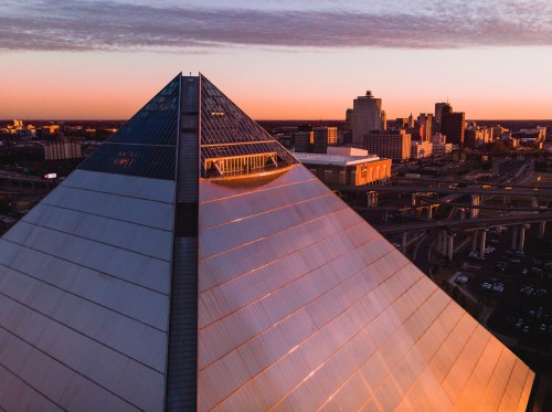 Aerial view of a pyramid structure at sunset, with a city skyline in the background and soft clouds in the sky.