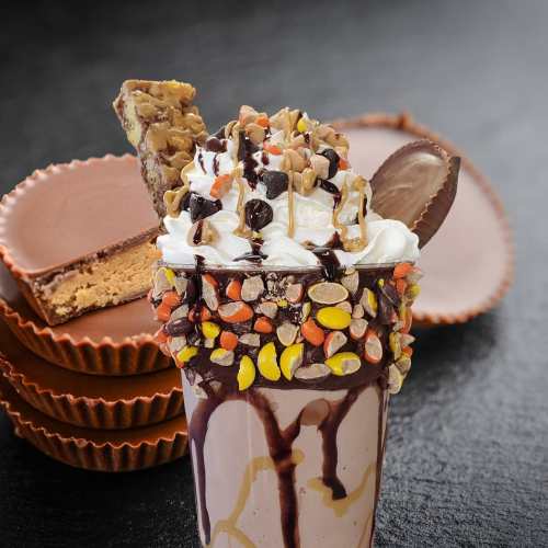 A decadent milkshake topped with whipped cream, chocolate drizzle, and candy, with chocolate cups in the background.