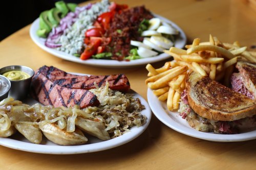 A table with two plates: one with grilled sausage, sauerkraut, and pierogies; the other with sandwiches and fries.