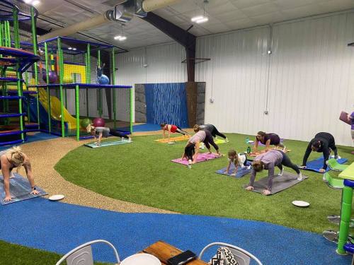 A group of adults and children practicing yoga on mats in a colorful indoor play area with slides and climbing structures.