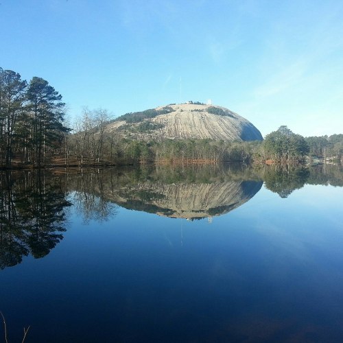 A serene lake reflects a large granite dome surrounded by trees under a clear blue sky.