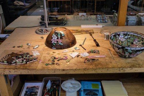 A workbench with a stained glass bowl, tools, and colorful glass pieces scattered around, illuminated by a lamp.