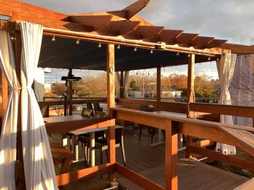 A wooden deck with curtains and string lights, featuring tables and chairs, set against a cloudy sky.