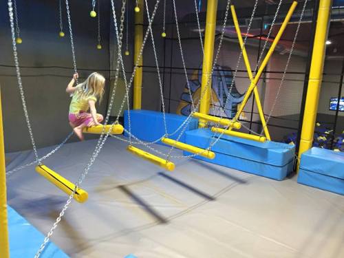 A child swings on a chain bridge in a colorful indoor play area with soft mats and climbing structures.