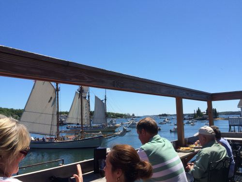 View of a harbor with sailing ships and boats, people dining at a waterfront restaurant under a clear blue sky.