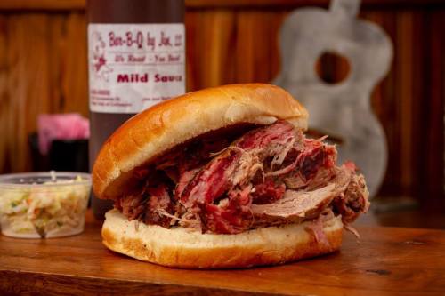 A close-up of a pulled pork sandwich on a bun, served with coleslaw and a bottle of mild barbecue sauce in the background.