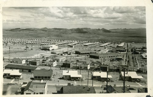 Aerial view of a mid-20th century town with rows of houses, streets, and distant mountains under a cloudy sky.