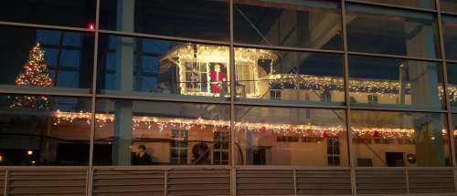 A festive scene reflected in a building's window, featuring Christmas lights, a tree, and a Santa figure.