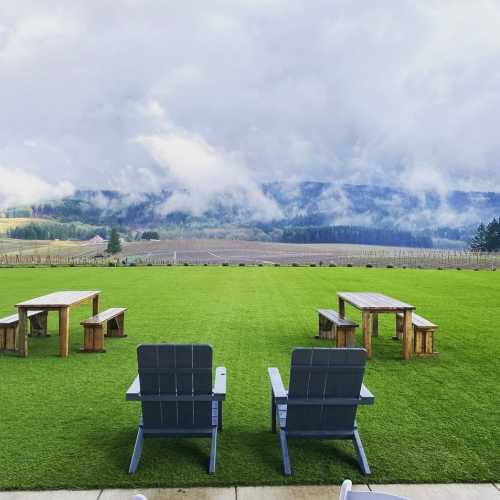 A scenic outdoor area with green grass, wooden tables, and chairs, set against a backdrop of cloudy hills.