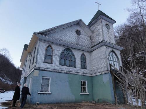 Two people stand beside an old, weathered church with a cross on top and arched windows, set against a mountainous backdrop.