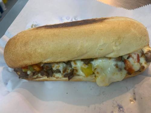 A toasted sub sandwich filled with beef, melted cheese, and colorful peppers, served on a white paper background.