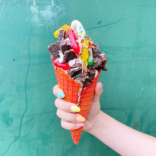A colorful ice cream cone topped with sprinkles, chocolate pieces, and syrup, held against a green background.
