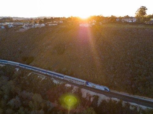 A train travels along a track beside a hillside at sunset, with houses visible in the background.