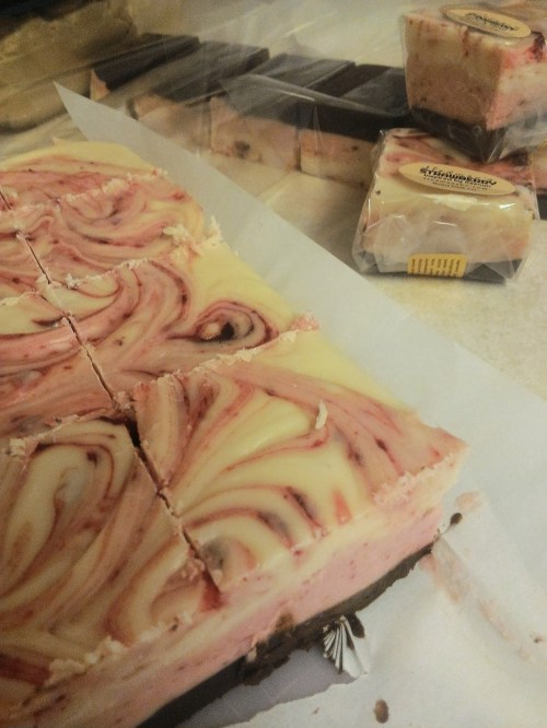 A close-up of swirled pink and white cheesecake bars on parchment paper, with packaged treats in the background.