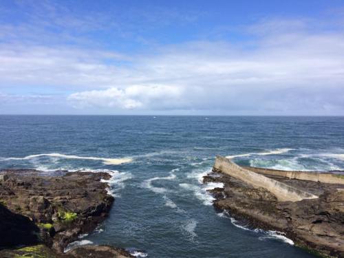 A rocky coastline meets a calm ocean under a blue sky with scattered clouds. Waves gently lap against the shore.
