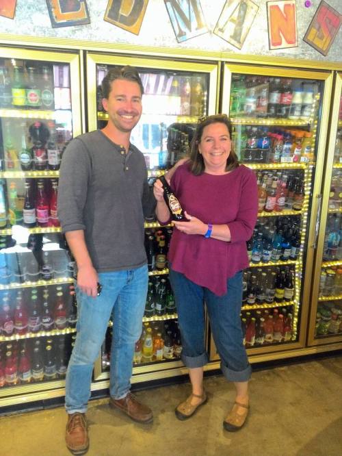 A man and woman stand in front of a colorful display of beverages, smiling and holding a bottle together.