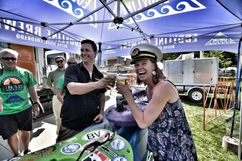Two people toast with drinks under a colorful tent, surrounded by smiling friends at an outdoor event.