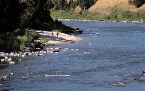 Two people walk along a sandy riverbank, with lush greenery and rocky shores beside a calm river.