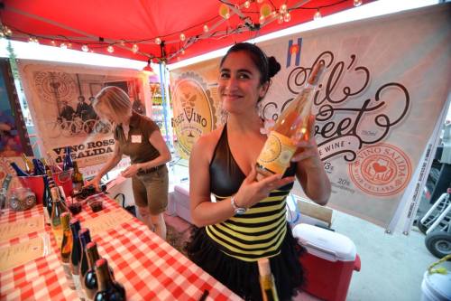A woman in a black and yellow dress holds a bottle of wine at a festive outdoor event with a red canopy.