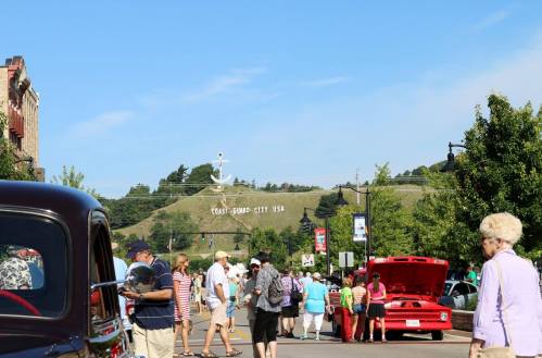 A busy street scene with people, classic cars, and a sign reading "Coast Guard City USA" in the background.