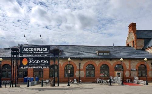 Exterior of a building featuring signs for Accomplice Beer Company and The Good Egg restaurant, with a cloudy sky above.