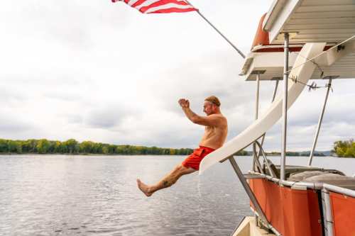 A man in red shorts slides off a boat into the water, with an American flag waving above him.