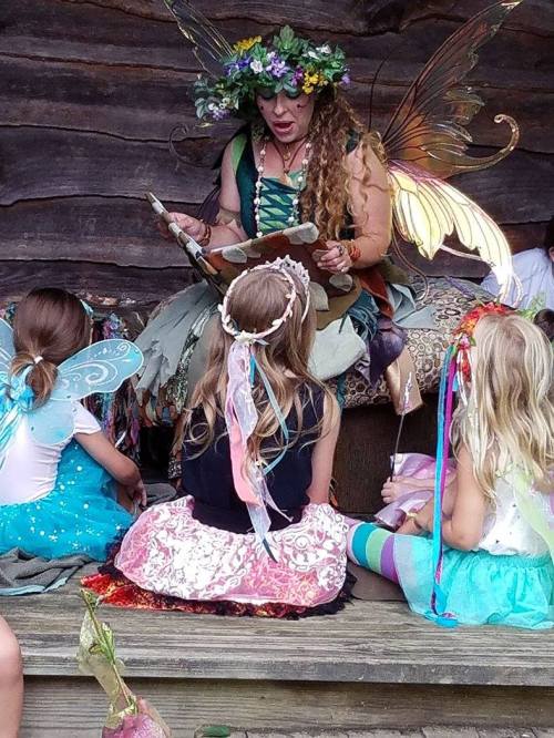 A fairy storyteller reads to children dressed in colorful costumes with wings, sitting on a wooden platform.