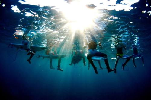 Underwater view of surfers sitting on boards, with sunlight streaming through the water's surface.