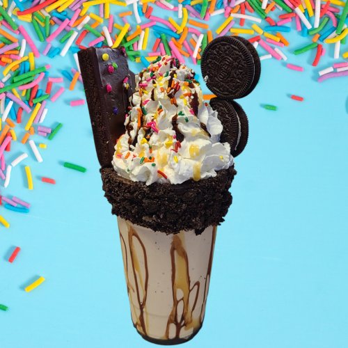 A decadent milkshake topped with whipped cream, chocolate, Oreos, and colorful sprinkles against a bright background.