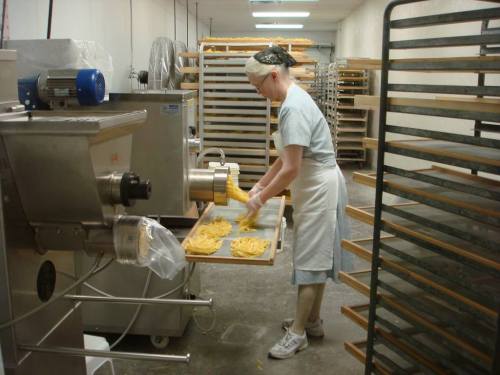 A woman in an apron prepares fresh pasta in a commercial kitchen, with racks and machinery in the background.