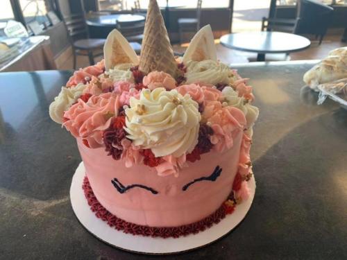 A pink unicorn cake decorated with flowers, a cone horn, and cute facial features, set on a table.