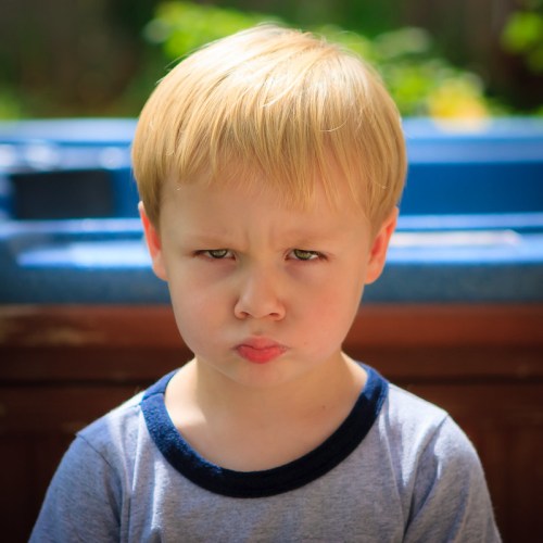 A young boy with blonde hair frowning, looking serious against a blurred outdoor background.