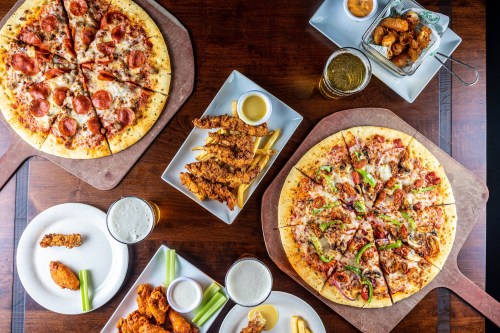 A table spread with pizzas, chicken tenders, dipping sauces, and drinks, showcasing a variety of delicious foods.