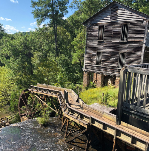 A rustic wooden mill with a waterwheel, surrounded by lush greenery and blue skies.