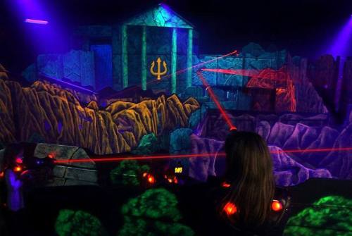 A person aims a laser gun in a colorful, illuminated laser tag arena with rocky terrain and a temple backdrop.