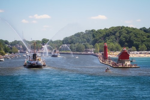 A scenic view of a harbor with boats, a lighthouse, and a crowd along the shore on a sunny day.
