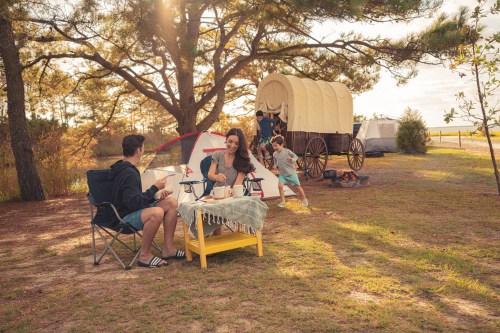 A family enjoys a camping trip with a tent and wagon, while a child plays nearby in a sunny outdoor setting.